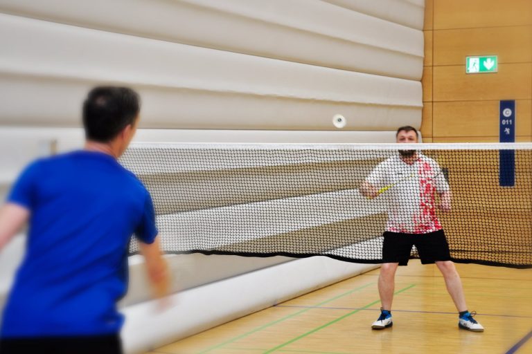 Zwei Spieler in einer Sporthalle spielen sich den Badmintonball über das Netz zu.