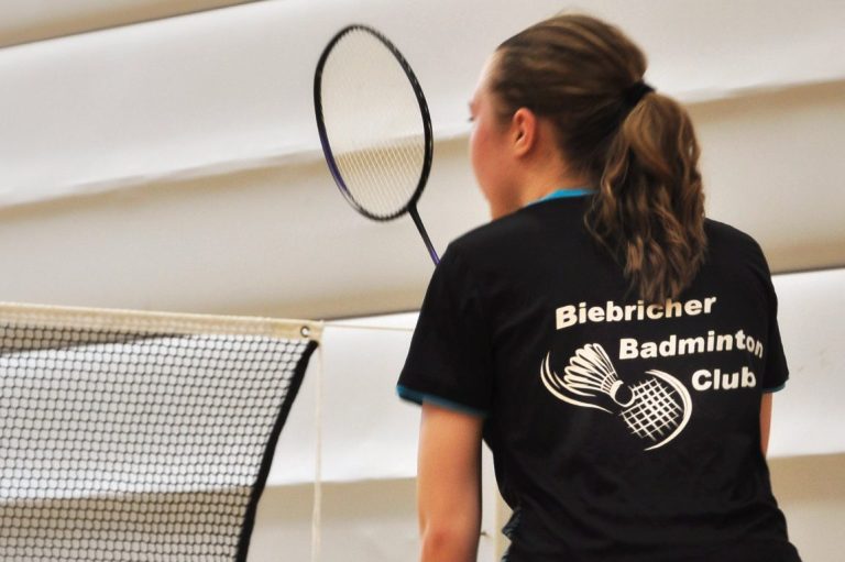 Junge Spielerin konzentriert mit Badmintonschläger am Netz in einer Sporthalle.
