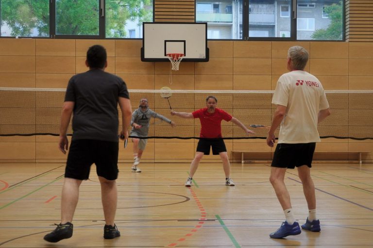 Vier Spieler mit Badmintonschlägern in einer Sporthalle spielen gegeneinander Badminton.